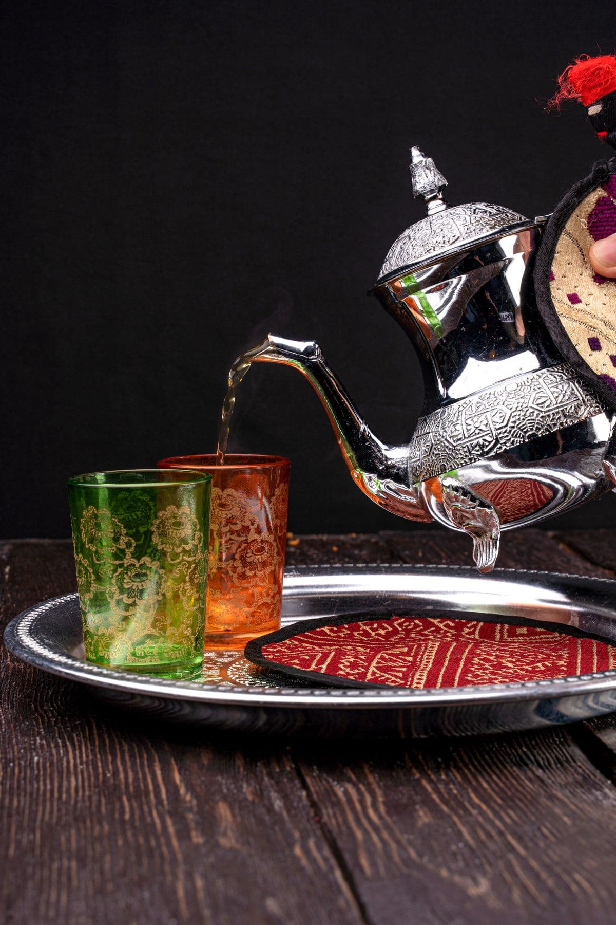 an ornate silver Moroccan teapot pouring golden mint tea in a high stream into decorated glasses on a tray